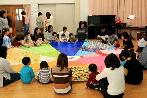 丸い大きな布を広げて、布の周りに縁になって布を引っ張りながら遊びを楽しむ親子