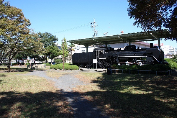 芝生や木々がある公園に蒸気機関車が展示されていて、その向こうにはブランコがある中央公園の写真
