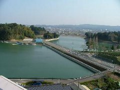 大きな貯水池の両端には山があり、奥には町並みが見える画像