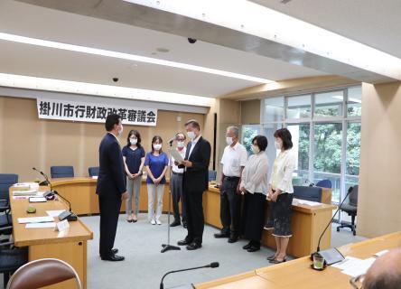 掛川市行財政改革審議会で手に書類を持った男性がマイクの前に立ち、それを取り囲むように立つ7名の参加者。