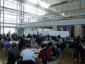 会場を後方から写した写真で、参加者がテーブルを囲んで座っている