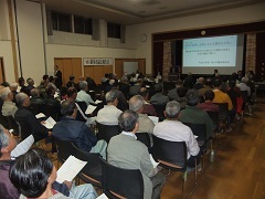 会場内に市民の皆さんが集まって椅子に座り前方でスクリーンに映しながら議会活動及び重要な課題について報告している様子