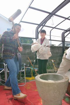 餅つきの様子。男性が臼を目掛けて杵を振り上げていて、近くには赤ちゃんを抱いた女性がその様子を見ている。