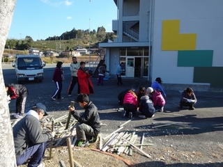 小学6年生と学校教職員と地域ボランティアとPTA理事らが竹を割ってジャンボ門松づくりをする様子